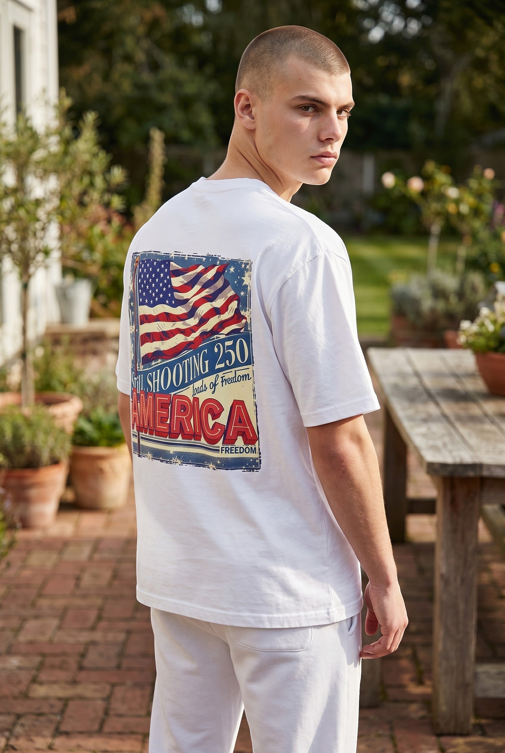 Naughty250 white heavyweight cotton t-shirt with distressed American flag graphic and "Still Shooting 250 America – Loads of Freedom" text on back.